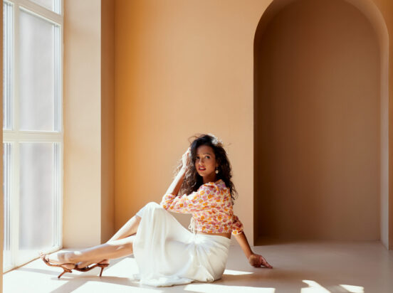 Charming young lady with dark hair in flower printed blouse, white long skirt and high heels sitting on the floor near big panoramic window with sunlight. Sexy model posing on camera in studio.