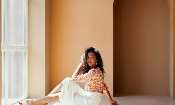 Charming young lady with dark hair in flower printed blouse, white long skirt and high heels sitting on the floor near big panoramic window with sunlight. Sexy model posing on camera in studio.