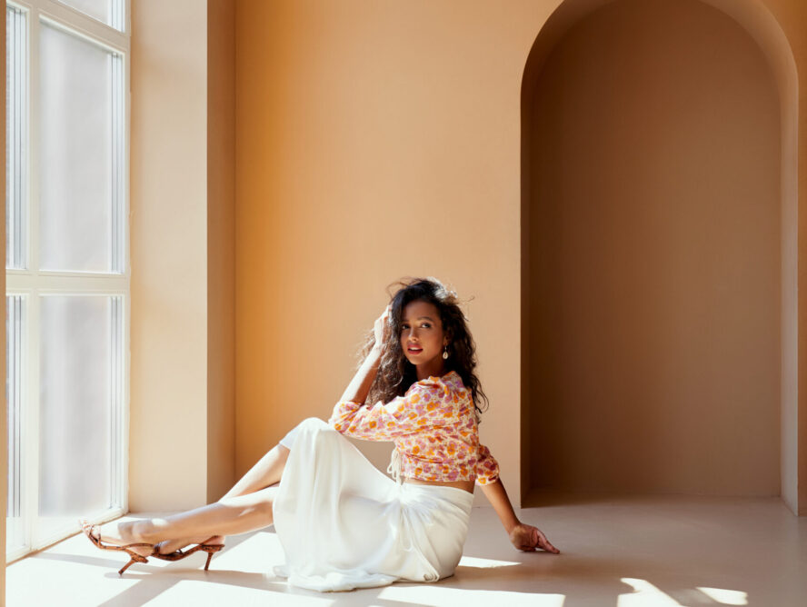 Charming young lady with dark hair in flower printed blouse, white long skirt and high heels sitting on the floor near big panoramic window with sunlight. Sexy model posing on camera in studio.