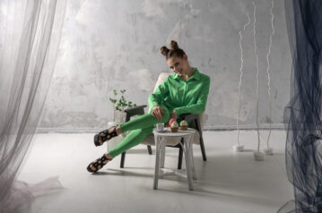 Cute girl smiles and drinks coffee with cupcakes in studio. Young woman in green suit poses on gray background.