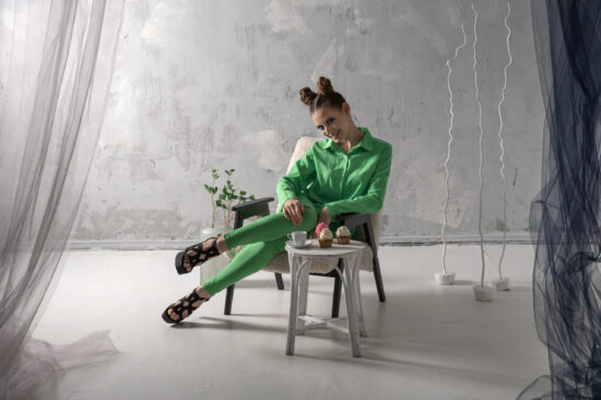 Cute girl smiles and drinks coffee with cupcakes in studio. Young woman in green suit poses on gray background.
