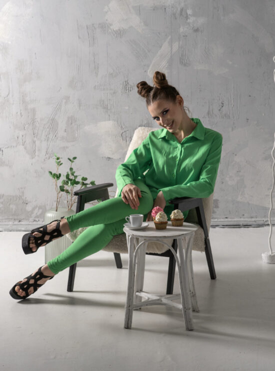 Cute girl smiles and drinks coffee with cupcakes in studio. Young woman in green suit poses on gray background.