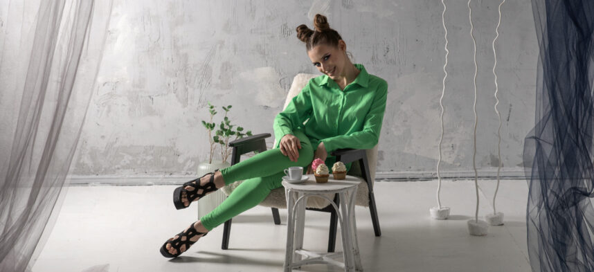 Cute girl smiles and drinks coffee with cupcakes in studio. Young woman in green suit poses on gray background.